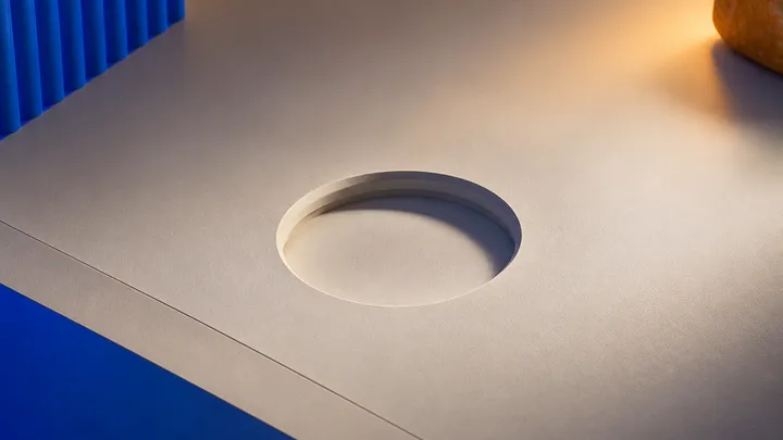 Un hueco circular vacío labrado en una superficie de escritorio, esperando un objeto redondo que no está, con un plano cobalto al fondo y una luz ámbar al costado.