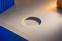 Un hueco circular vacío labrado en una superficie de escritorio, esperando un objeto redondo que no está, con un plano cobalto al fondo y una luz ámbar al costado.
