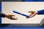 Dos manos abiertas entregándose un testigo de relevo azul cobalto con una franja ámbar, frente a un panel rectangular claro y vacío.