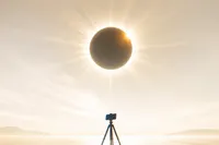 Ilustración editorial de un eclipse solar total bajo en el horizonte, con la silueta de un teléfono en trípode en primer plano