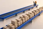 Dos cintas transportadoras azul cobalto en paralelo: una cargada de estibas con cajas de cartón idénticas y la otra completamente vacía salvo por una sola pieza metálica plegada, de interior ámbar, al final del recorrido