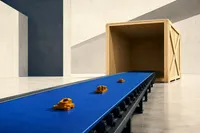 Una cinta transportadora azul cobalto que lleva apenas tres piezas ámbar hacia un enorme cajón de embalaje de madera vacío