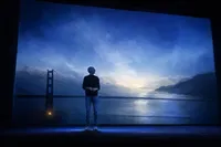 Figura anónima en silueta sobre un escenario oscuro bajo un haz de luz, con una nube que se deshace en partículas hacia arriba.
