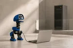 Un pequeño robot bípedo azul de un ojo y pico plano, unido por un cable corto a un portátil plateado abierto, mientras al fondo un armario de servidores queda tras una mampara de vidrio que ningún cable cruza.