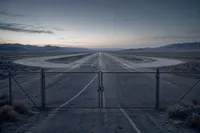 Una pista de pruebas vacía en el desierto, con una reja cerrada en primer plano y una luz ámbar tenue perdiéndose en el horizonte.